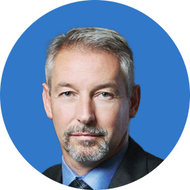Un homme blanc sur un fond bleu portant une chemise bleue et une veste noire. Il a les cheveux blancs.